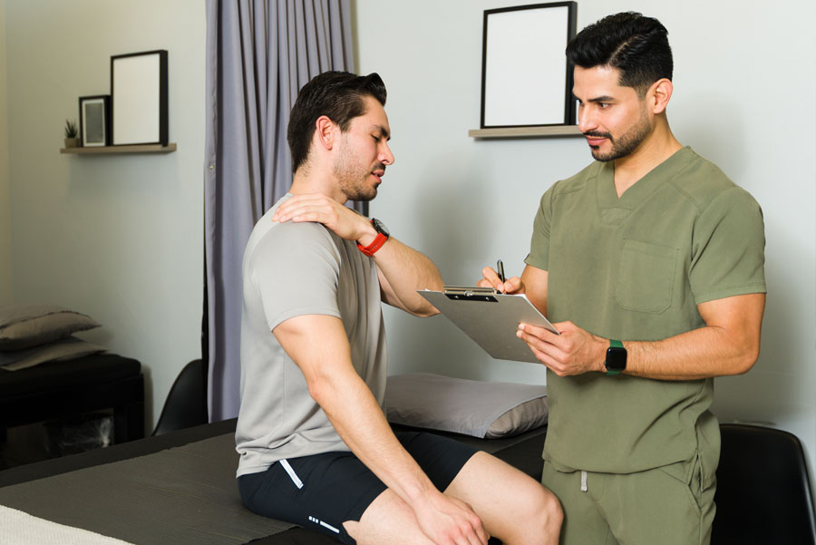 Male doctor or nurse with clipboard in medical office speaking with male patient who’s touching his shoulder after an injury