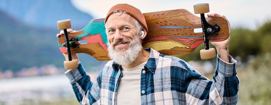 Happy, bearded, elderly hipster man standing in nature park holding skateboard