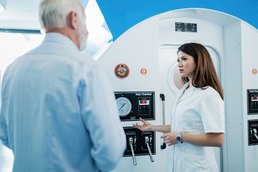 Female doctor talking to elderly male patient in front of hyperbaric oxygen therapy chamber at clinic
