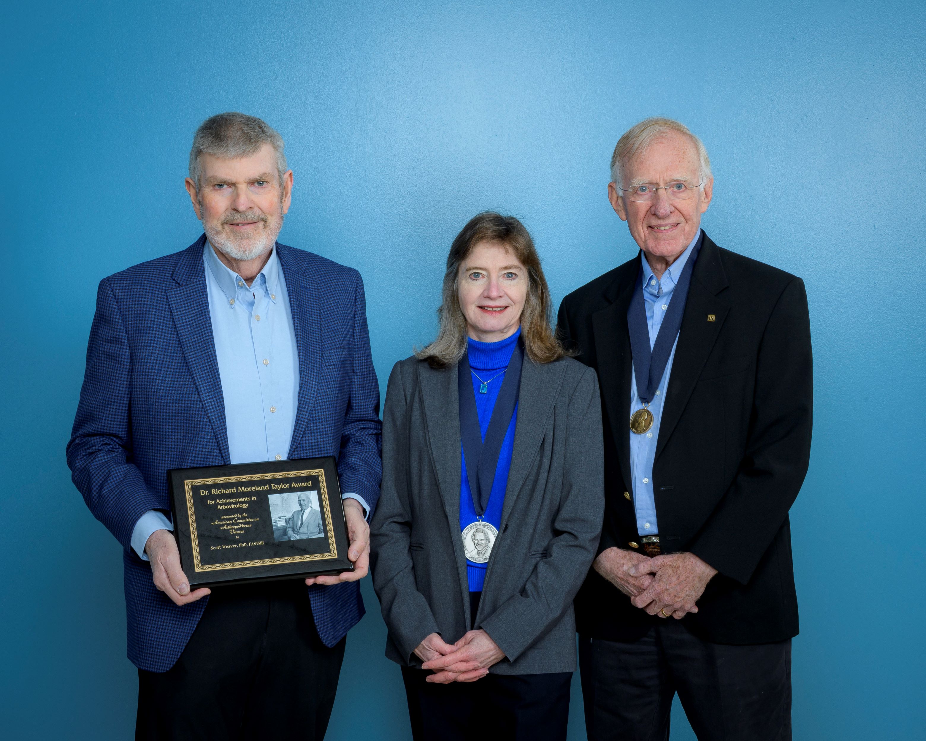 Three individuals stand indoors against a neutral background, with the individual on the left holding a framed plaque while the other two stand beside them. All are dressed in professional attire.