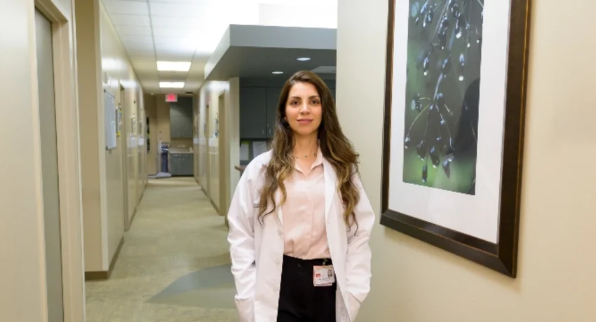 A person wearing a white lab coat stands in a well-lit medical office hallway. Framed artwork hangs on the wall beside the individual, and exam room doors and clinical workspaces are visible in the background.