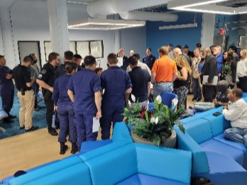 Large group of staff gathered in a modern operations room, standing in a circle near blue seating while participating in a group discussion.