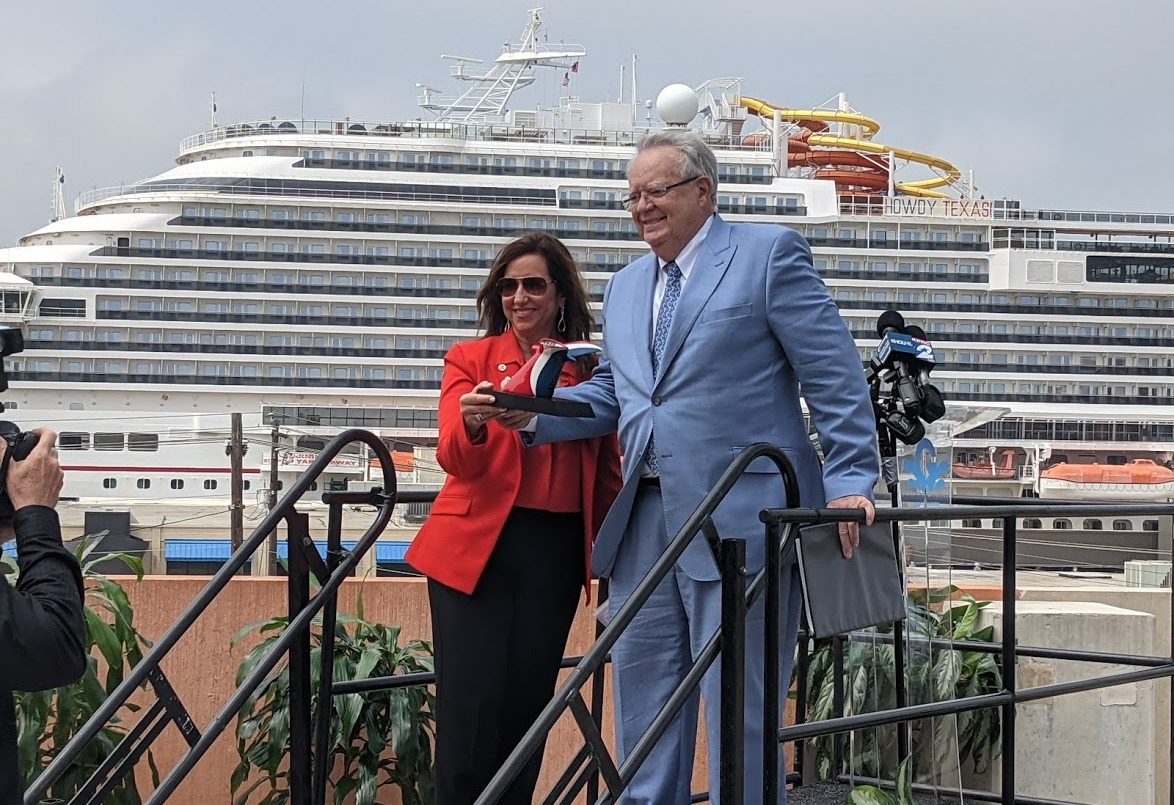 Two officials stand on an outdoor platform in front of a large cruise ship while holding a ceremonial item, with media equipment nearby.