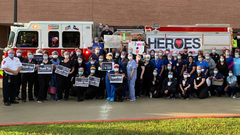 Large group of masked staff and responders standing in front of a fire truck, holding signs and banners expressing appreciation.
