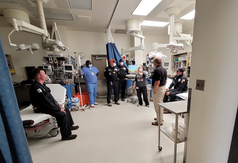 Medical staff and emergency personnel stand in a hospital treatment room surrounded by equipment during a group briefing.