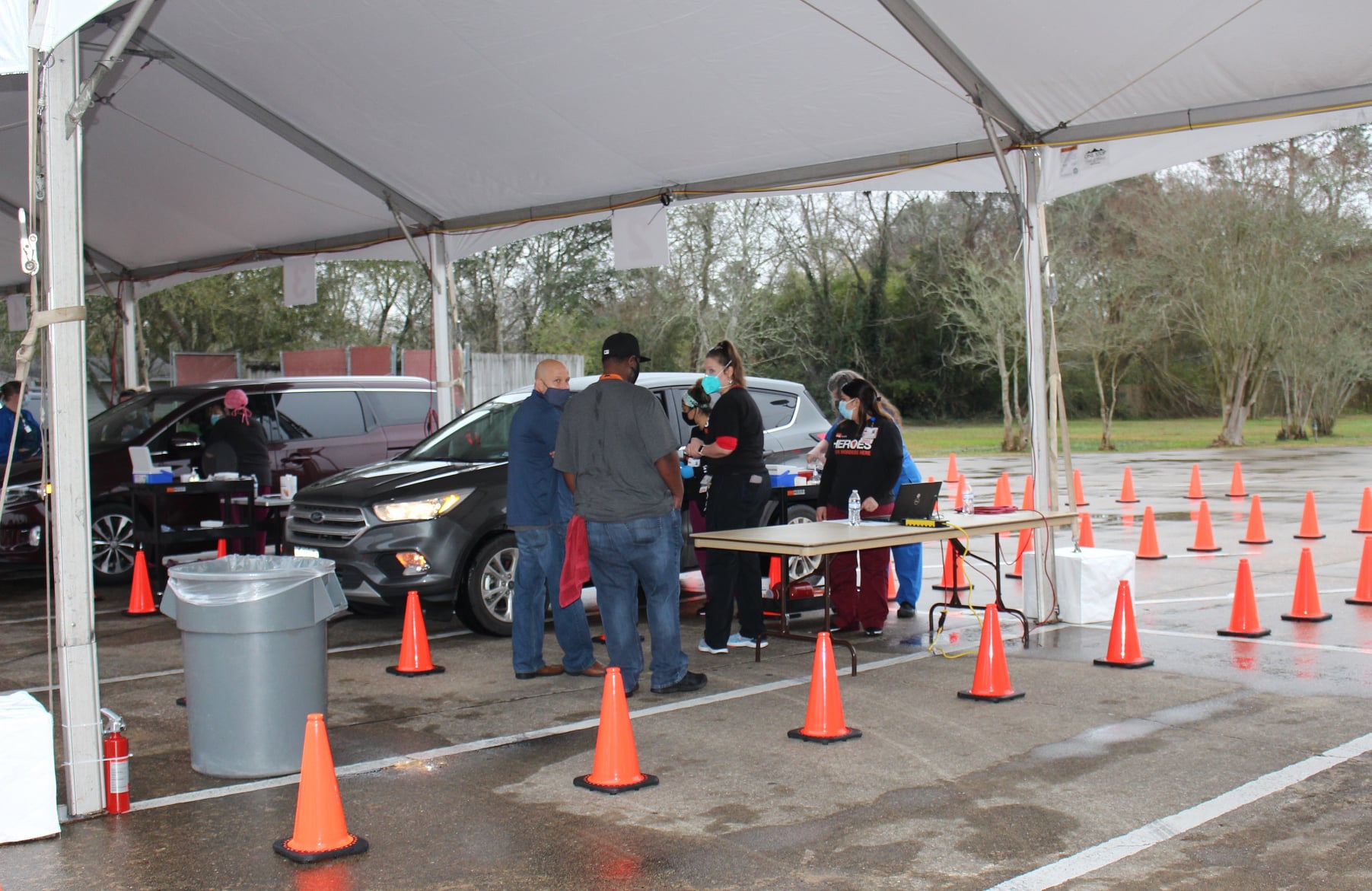 Vehicles pass through a covered outdoor screening station with traffic cones while staff assist individuals at a table and near cars.