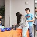 Students enter a room while one individual signs in at a table with colorful bowls and materials, and another person stands nearby assisting with the check-in process.