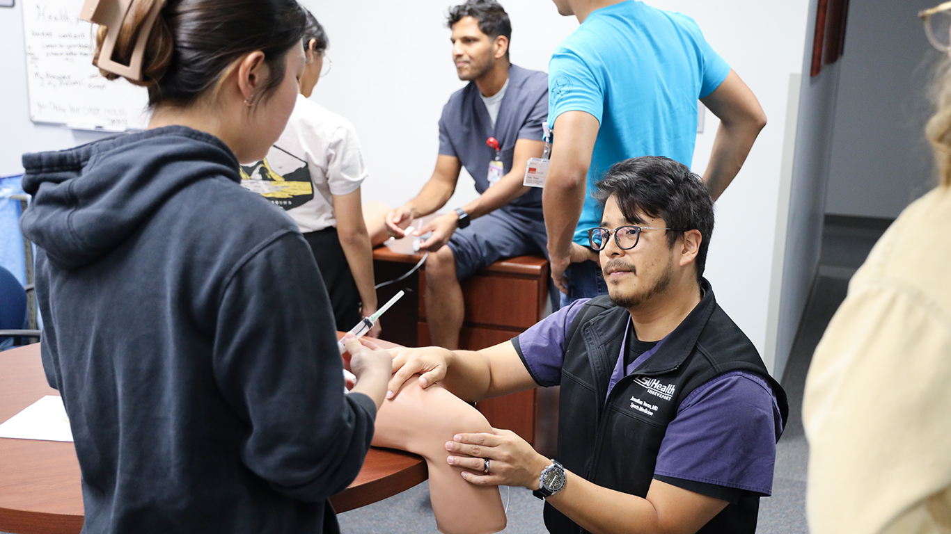 Students seated around a table practicing patient assessment skills on a classmate’s leg during a clinical training session.