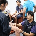 Students seated around a table practicing patient assessment skills on a classmate’s leg during a clinical training session.
