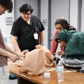 Medical students practice injection techniques on simulation models at a table while an instructor observes and guides in a clinical training room.