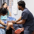 Healthcare trainees in a clinical room practicing a knee examination on a seated patient, with medical equipment and supplies visible in the background.