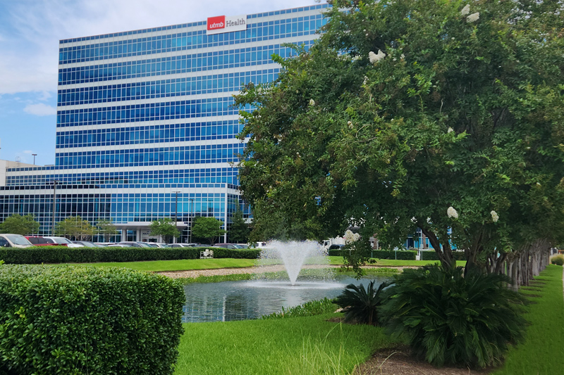 Exterior view of a UTMB Health building with a glass facade, landscaped lawn, and water fountain.