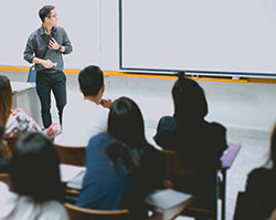 An Asian man with glasses stands in front of a classroom of adult students, pointing a laser pointer at a white board