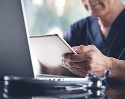 A man in dark blue scrubs holds a computer tablet while seated at a desk with a laptop and stethoscope on it.