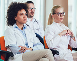 A black woman, a white woman, and a white man sitting in white lab coats and sitting in lecture hall chairs in a brightly lit room looking off camera
