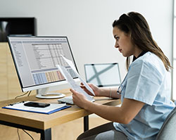A white woman in light blue scrubs sitting at a desk with a large computer monitor and laptop, looking at papers.