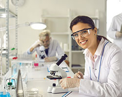 A white woman wearing a lab coat and safety googles is looking at the camera and smiling while her hands are on a small desktop microscope.