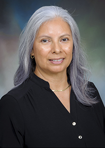 Formal studio portrait of an individual with long gray hair wearing a black top, positioned against a softly blended blue and green background.