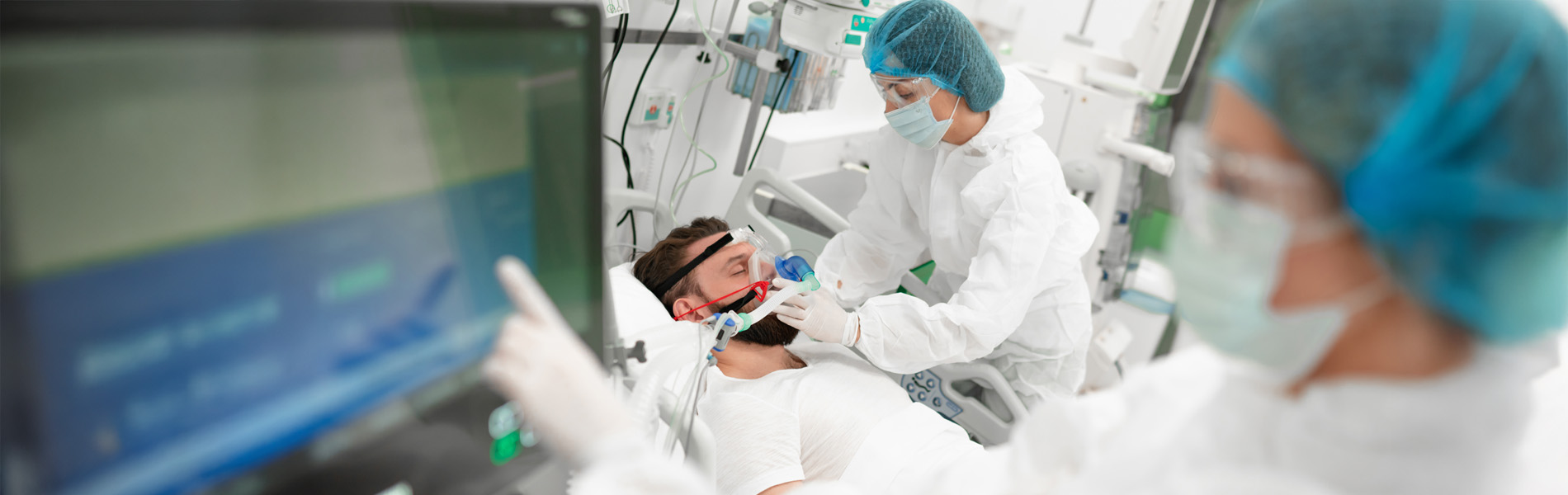 Healthcare worker in full protective gear adjusts breathing mask on male patient in hospital bed, surrounded by medical equipment, highlighting critical care, respiratory support, patient treatment
