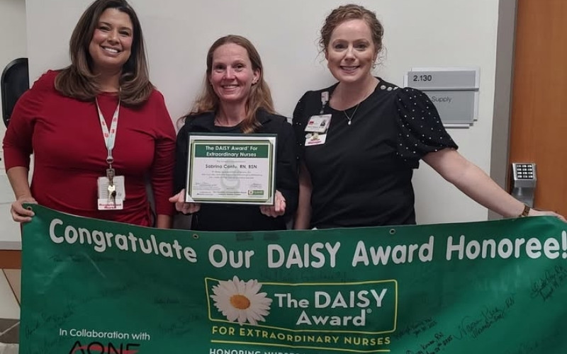 A DAISY Recipient poses with her certificate and two colleagues behind a DAISY Award banner.