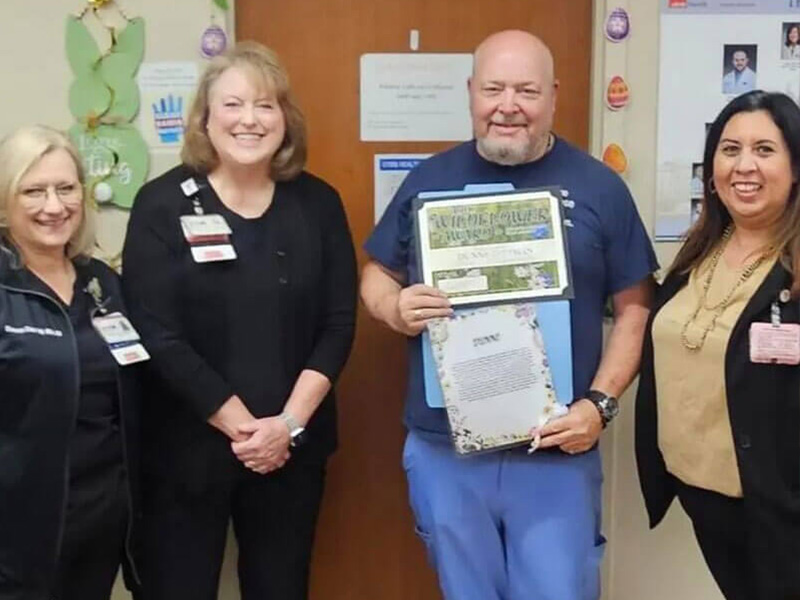Wildflower recipient, Dunne Dittman, smiles holding his nomination letter and award certificate surrounded by members of the Nursing Services team.