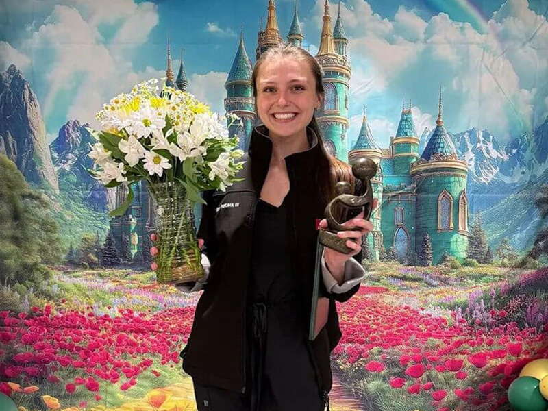 DAISY Award recipient, Elissa Peipleman, smiles holding her award and a bouquet of daisies in front of a Wizard of Oz themed backdrop.