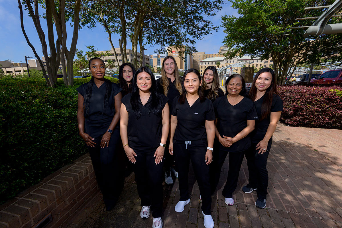 A group of smiling nurses in black scrubs stand together for a group portrait in a garden.