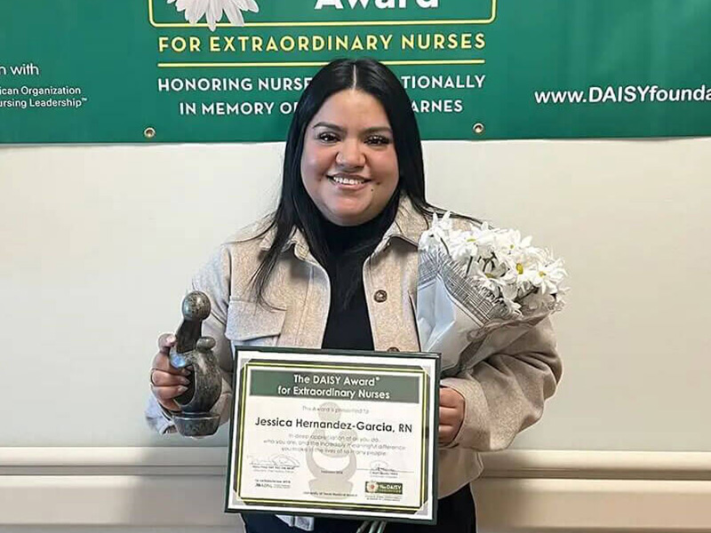 DAISY Award recipient, Jessica Hernandez-Garcia, stands smiling holding her award, certificate and a bouquet of daisies.
