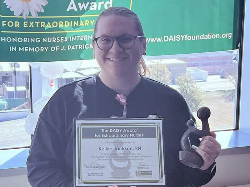 DAISY Award recipient, Kellye Jackson, stands smiling holding her certificate and award.