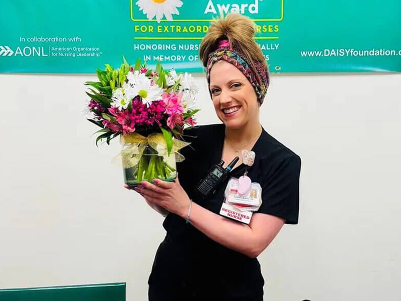 DAISY Award recipient, Krista Avants, smiles holding colorful flowers in front of the DAISY Award banner.