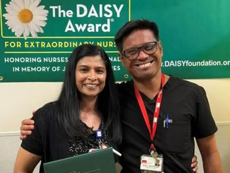 DAISY Award recipient Nissy George smiles with her certificate next to another nurse in black scrubs.