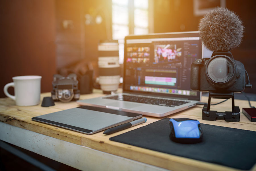 A digital camera, lenses, and a laptop with video editing software are arranged on a workstation for UTMB media production.