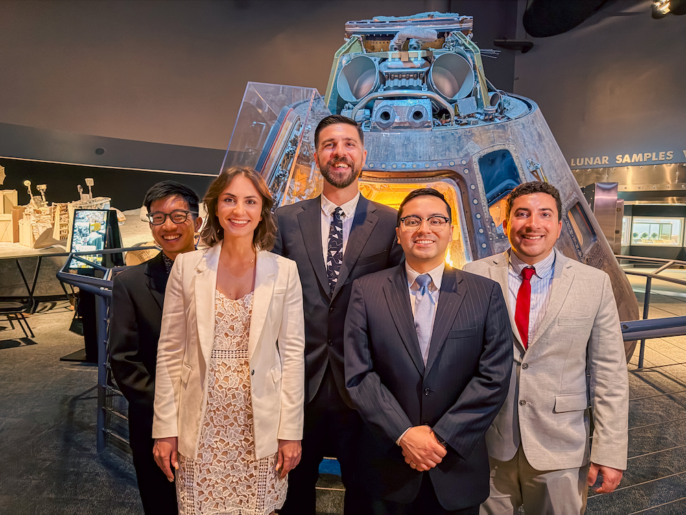 Five UTMB Aerospace Medicine Residency graduates stand together in formal attire in front of a spacecraft capsule exhibit at Space Center Houston’s Starship Gallery.