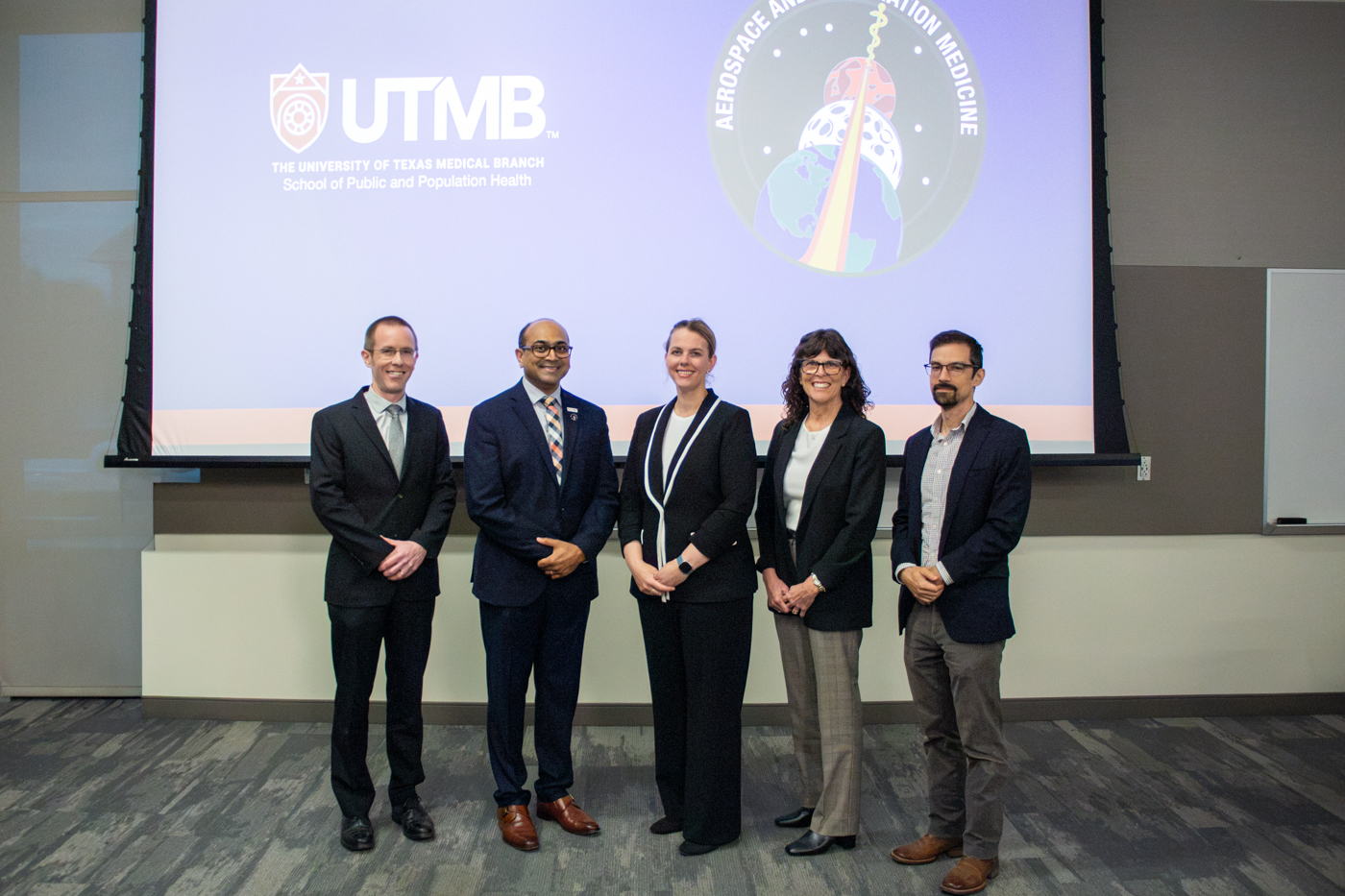 From left, Drs. Matthew Makowski, Ronak Shah, Amy Kreykes, Mary Van Baalen, and John Marshall stand beneath the Aerospace and Exploration Medicine patch on a screen.