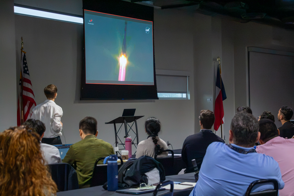 PASM participants in a conference room at Space Center Houston watch a projection screen showing a Soyuz rocket lifting off, with U.S. and Texas flags flanking the screen.