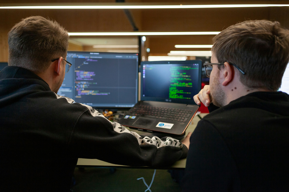 Two students collaborate at a workstation, reviewing computer code displayed on a laptop and external monitor in a modern research and technology workspace.