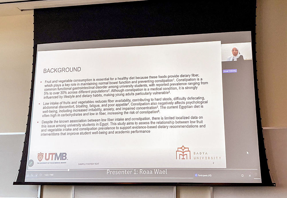 Projected slide from a Badya University student presentation shown on a large screen during the poster session, with the presenter visible in a small video window.