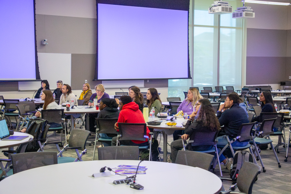 Students, faculty, and staff sit at round tables in a UTMB classroom while listening to the Community Voices Panel during National Public Health Week.