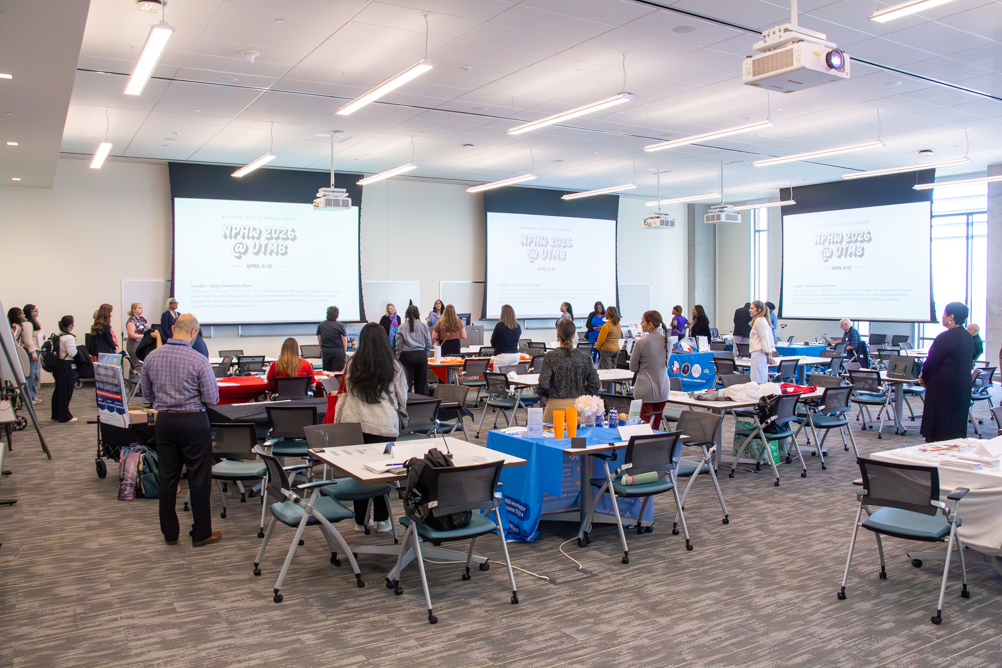 Wide view of the UTMB National Public Health Week Career Connections Mixer, with students and partner representatives gathered around tables in a large conference room beneath projection screens showing the event schedule.