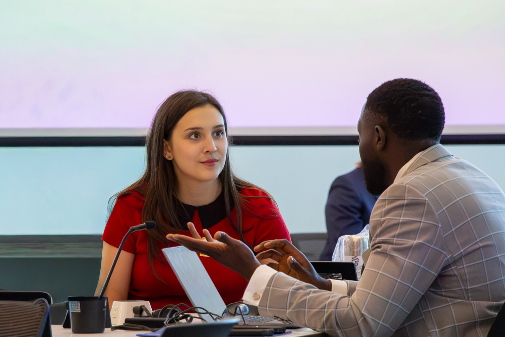 A student in a red top speaks across a table with a partner representative in a gray suit during a one-on-one networking conversation.