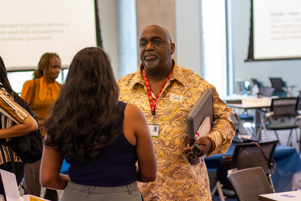 A student in a patterned shirt holds folders while speaking with a partner during a standing conversation at the mixer.