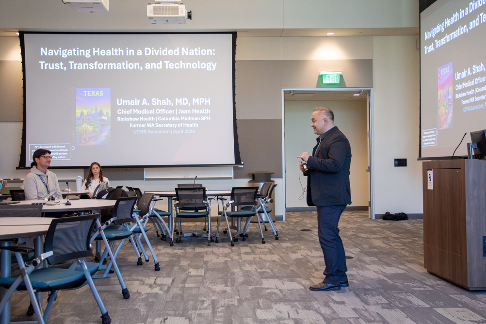 Speaker presents in a classroom-style event space during a keynote talk, with the presentation title displayed on large screens behind him.