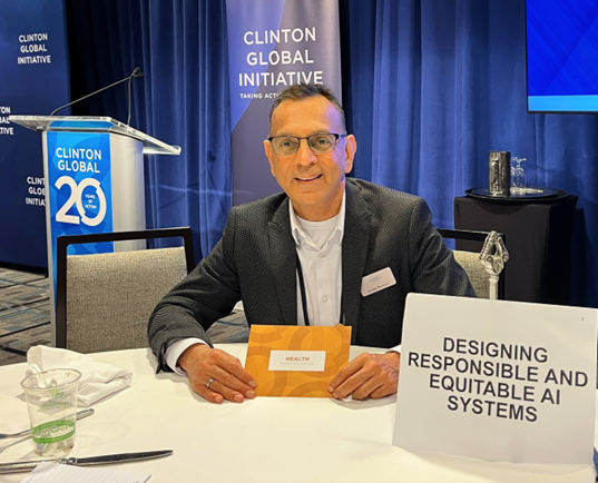 Dr. Suresh Bhavnani seated at a Clinton Global Initiative event table with a sign reading “Designing Responsible and Equitable AI Systems.”