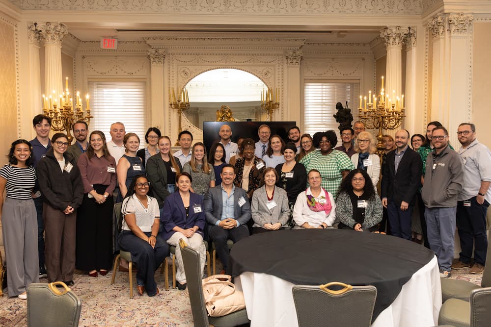 A large group of symposium attendees gathered for a group portrait, standing and seated around a covered round table in an ornate room with chandeliers.