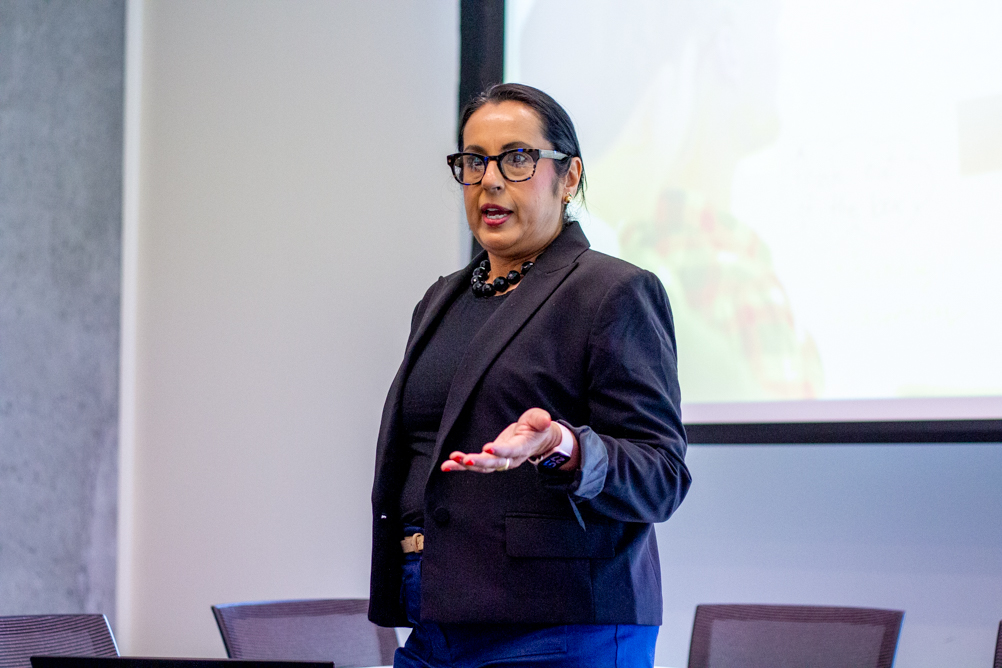 Dr. Monisha Arya in a black blazer stands at the front of a room giving a presentation, gesturing with one hand while speaking; a projected slide is visible behind her.