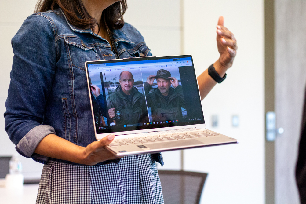 A person wearing a denim jacket holds an open laptop showing two side-by-side photos of a man in outdoor gear, while gesturing with their other hand during a presentation.