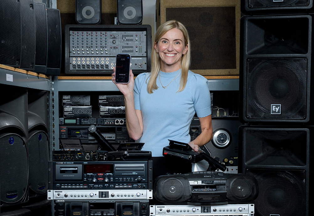Dr. Meredith Masel smiles and holds up a smartphone in front of a wall of vintage cassette decks, mixers, and speakers, a portable tape recorder in her other hand.