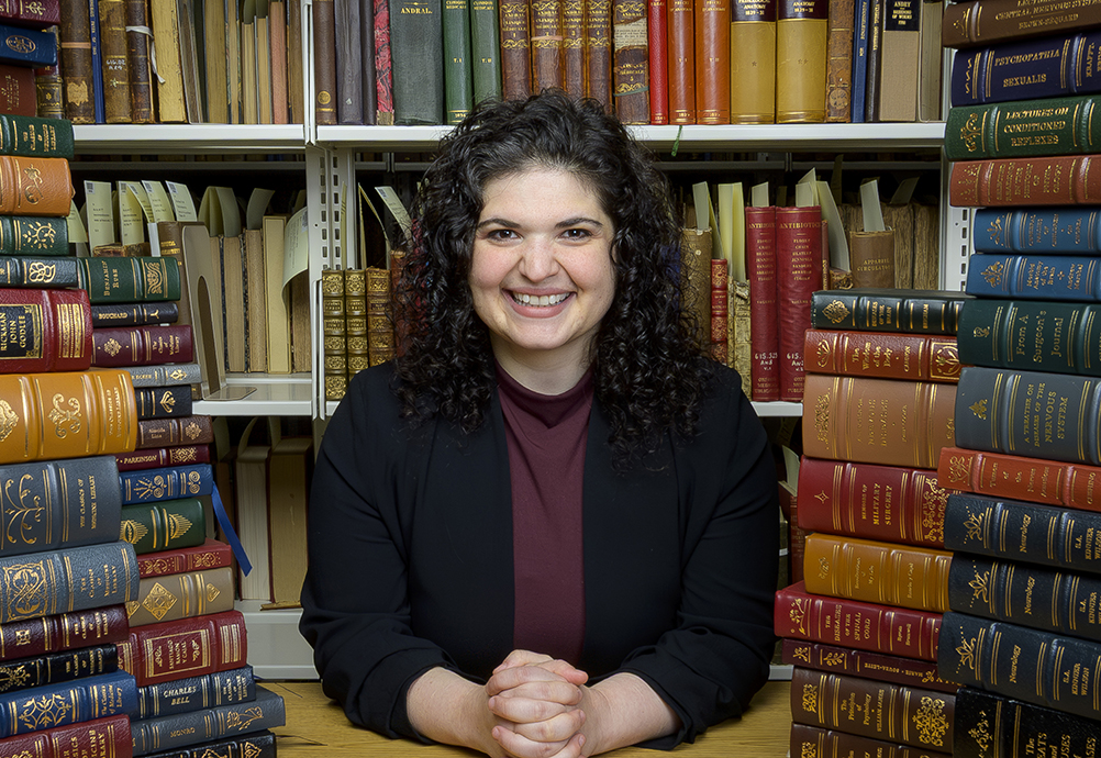 Dr. Rich smiles while seated at a table between stacks of historical medical books in a library or archive setting.