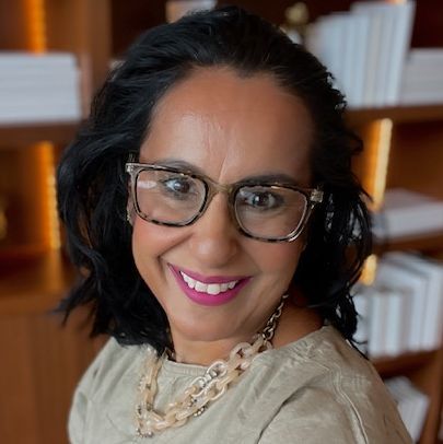 Professional headshot of Dr. Monisha Arya with shoulder-length dark hair and glasses, smiling and looking slightly to the side, wearing a light-colored top and necklace against a softly lit indoor background.
