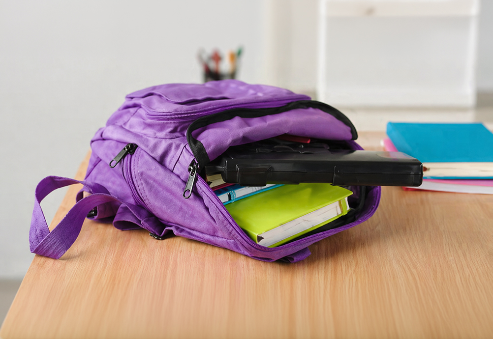 Open purple backpack on a wooden desk with school supplies inside, including notebooks and a black case, with additional folders and books blurred in the background.
